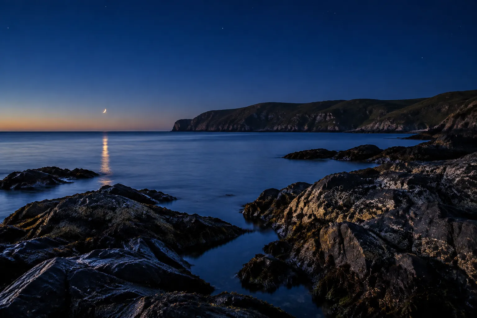 A rocky coastline at blue hour with a calm sea, a far headland and a thin crescent Moon low over the water.