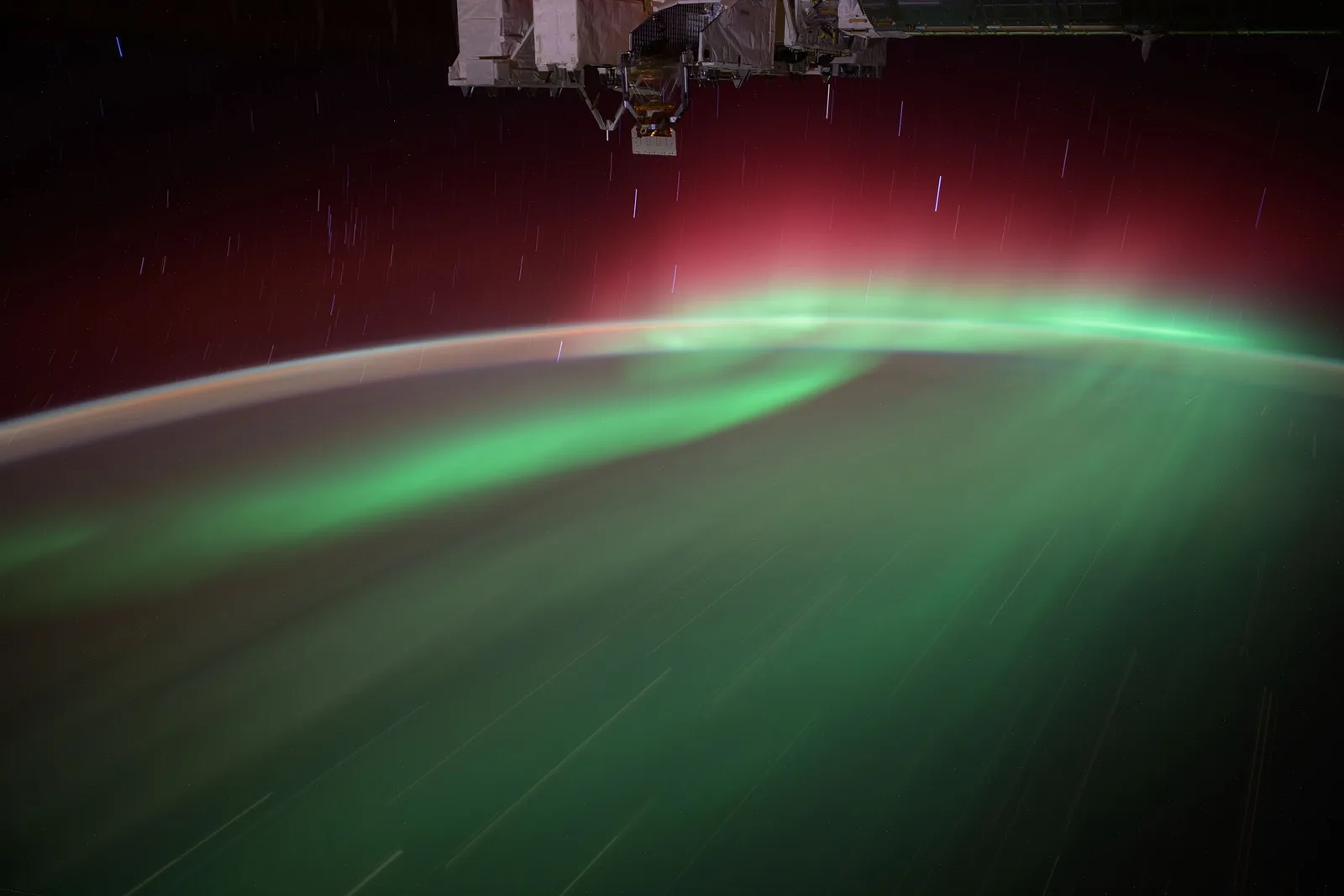 A green aurora with a red upper fringe streaking along Earth’s curved horizon, seen from the International Space Station.