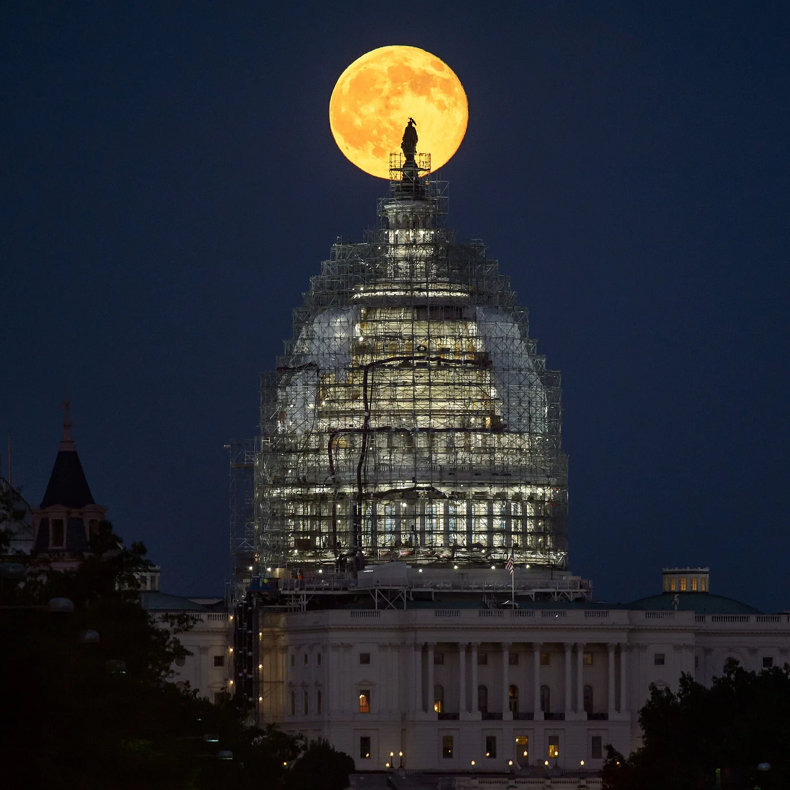 A full Moon rising directly behind the scaffolded dome of the US Capitol.