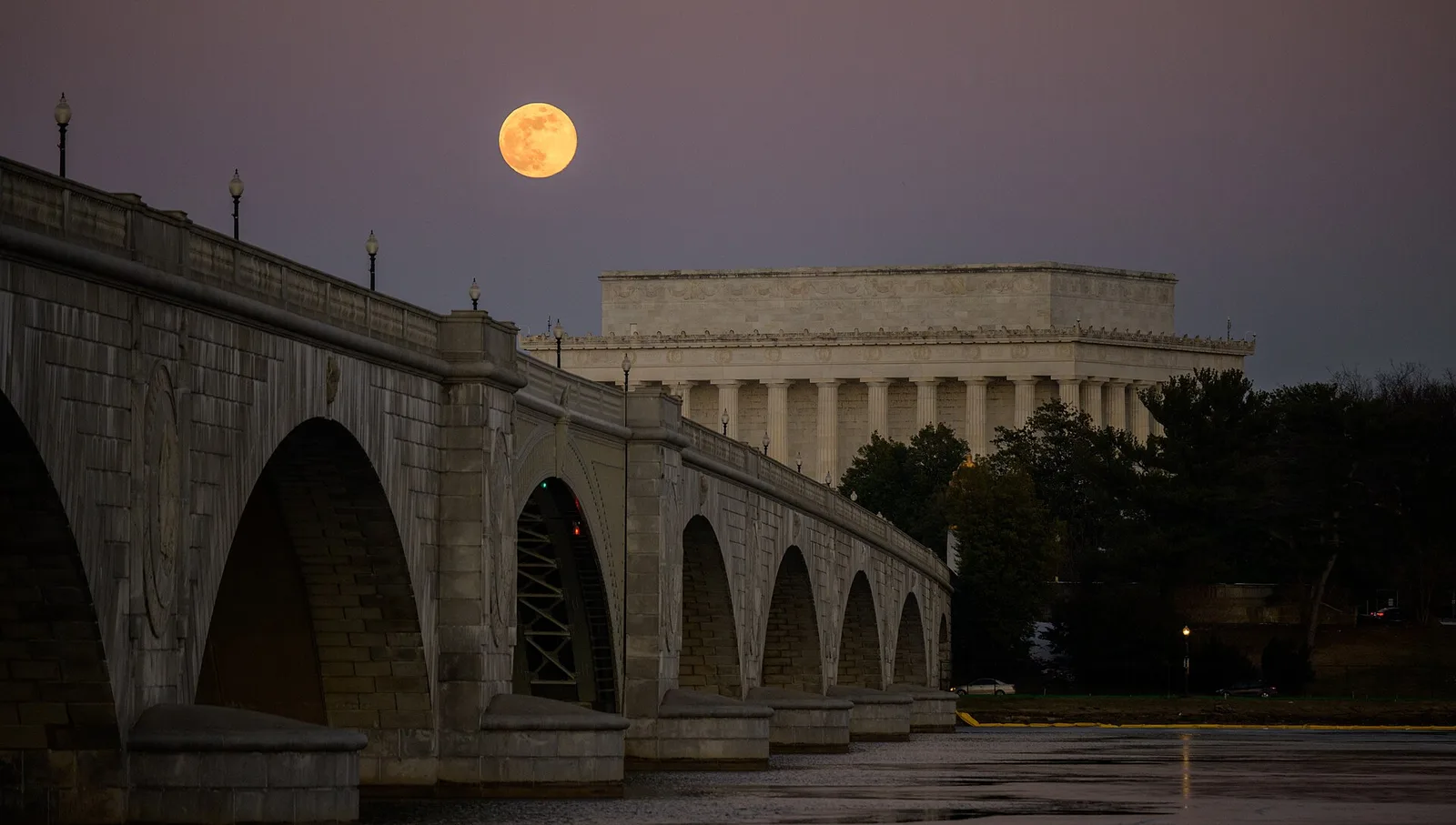 A full Moon rising beside a stone bridge and the Lincoln Memorial at dusk.