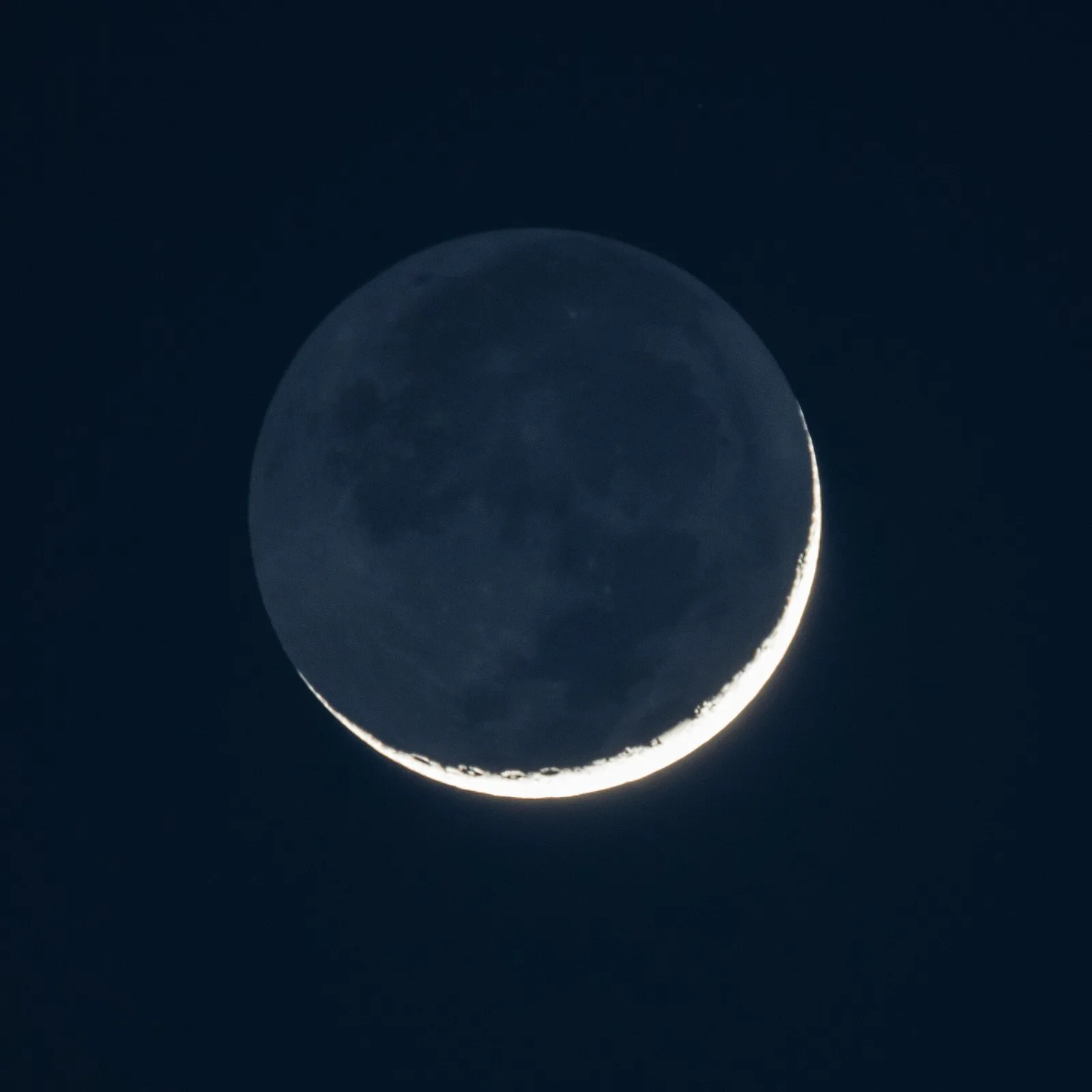 A very thin crescent Moon with its dark part faintly lit.