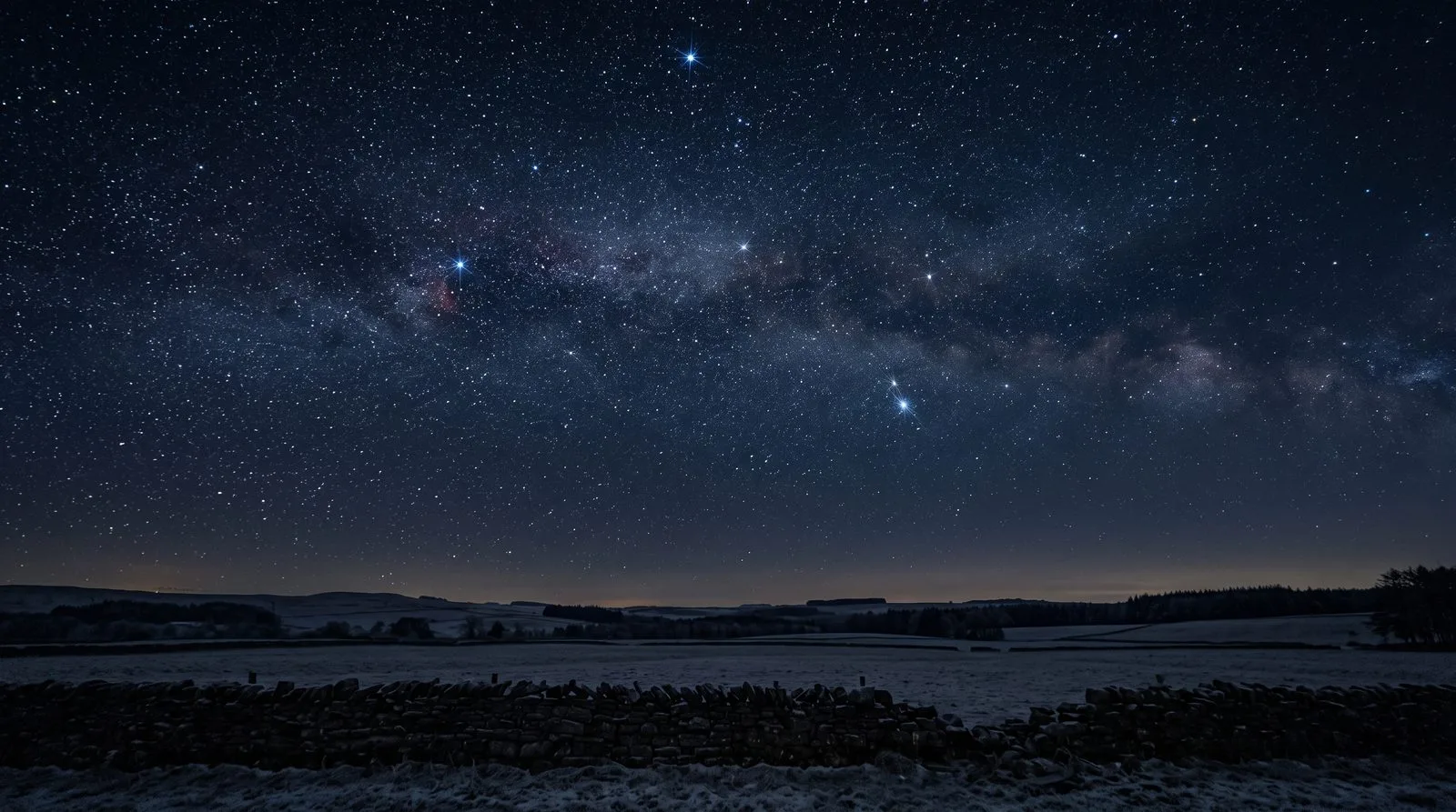 The Milky Way arching over a dark-sky landscape, a drystone wall and frosted fields in the foreground under a horizon glow.