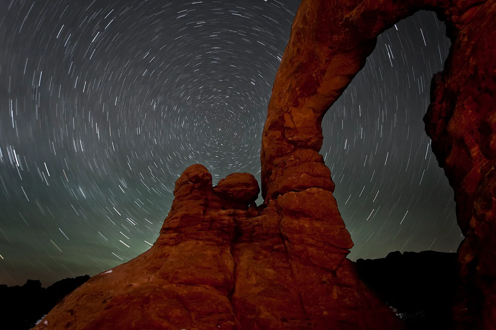 Star trails circling one point above a red rock arch.