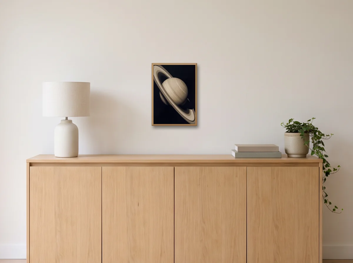 Saturn as an A3 poster in a natural wood frame on an off-white wall above an oak sideboard with a lamp and plant