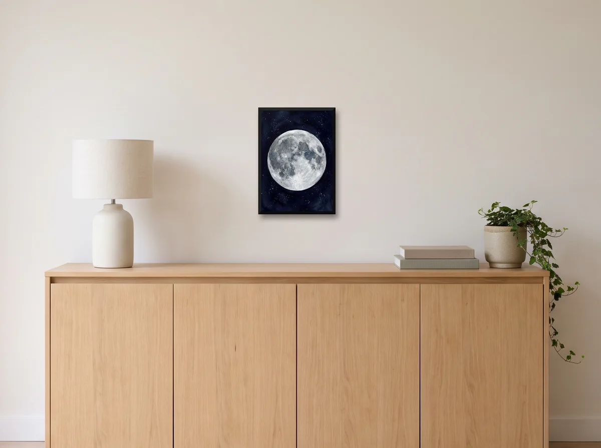 Watercolour Moon as an A3 poster in a black frame on an off-white wall above an oak sideboard with a lamp and plant
